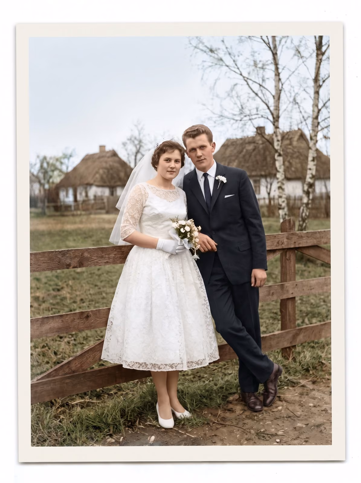 After: Bride and groom leaning on a wooden fence in front of village houses, restored from a faded black and white print
